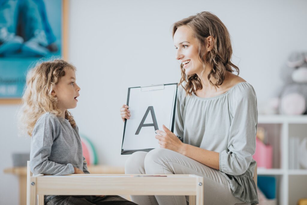 Uma menina praticando a pronunúncia com uma mulher segurando um quadro com a letra "A" escrita nele.